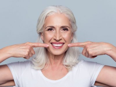 Concept of having strong healthy straight white teeth at old age. Close up portrait of happy with beaming smile female pensioner pointing on her perfect clear white teeth, isolated on gray background