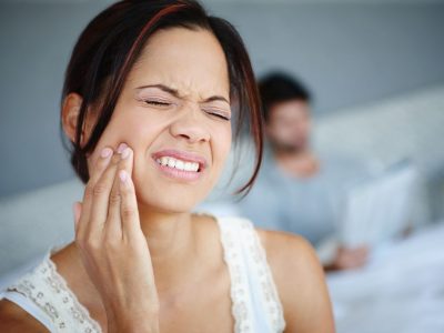 Shot of a woman sitting on the side of her bed with bad toothache with her boyfriend in the background